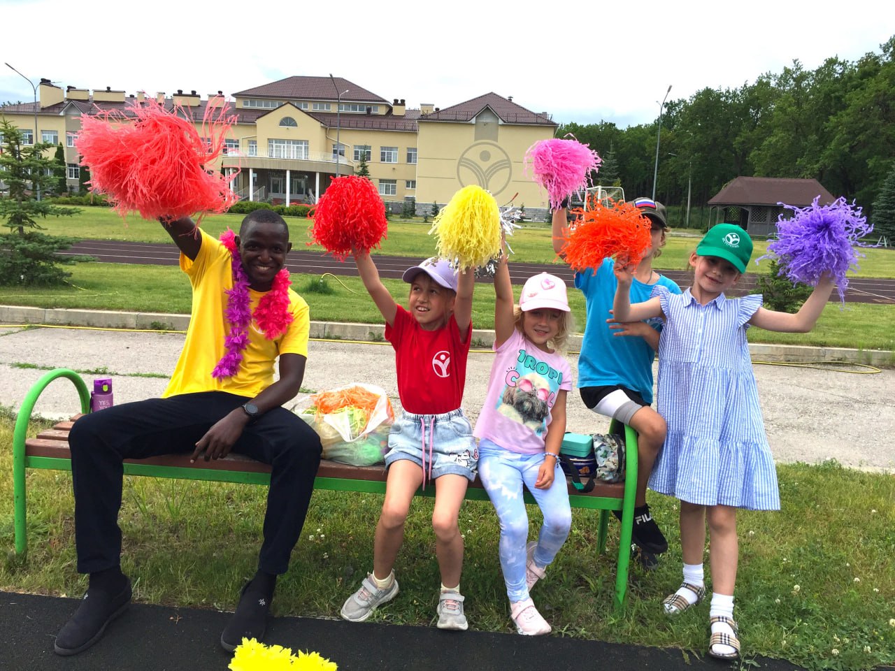 Children with brightly colored pom-poms sit on a bench at the camp