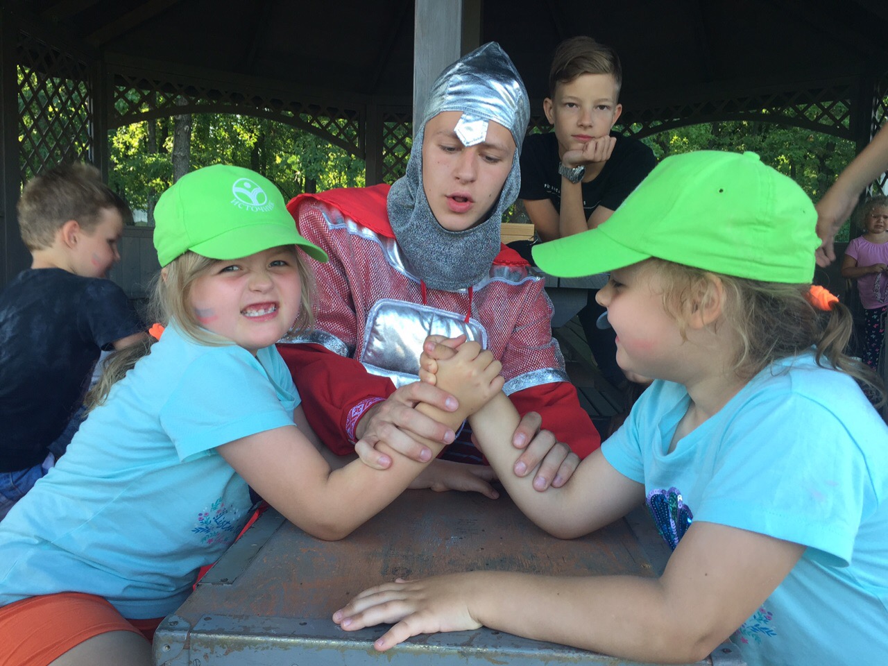 Children in summer camp participate in arm wrestling under the supervision of a counselor in a knight costume