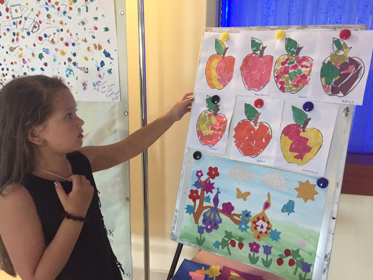 Girl presenting crafts of bright apples and floral collage at camp exhibition