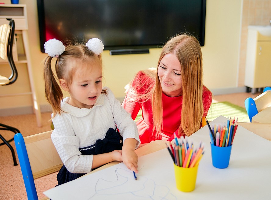 Little girl drawing at the table with her teacher in kindergarten
